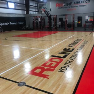 Indoor basketball court with red line athletics logo and weight lifting area.